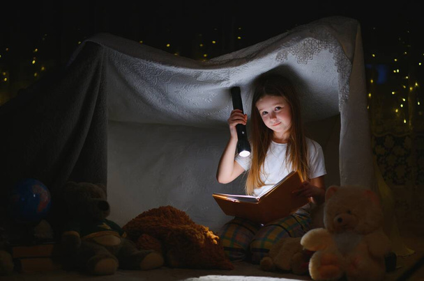 child girl reading with book and flashlight and teddy bear in tent. before going to bed - Fotografie, Obrázek