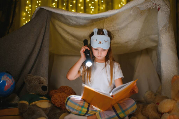 happy child girl laughing and reading book in dark in a tent at home - 写真・画像