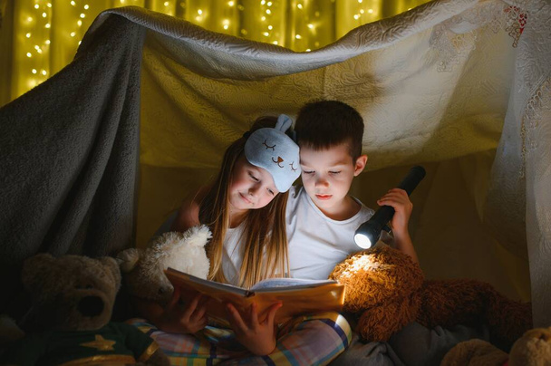 Two children with flashlight read a book under a blanket as a tent - Φωτογραφία, εικόνα