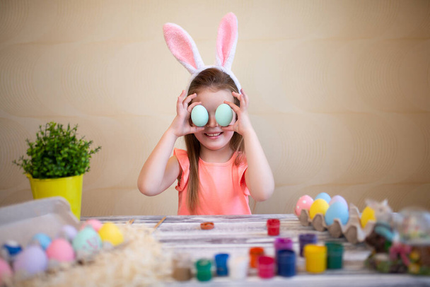Happy little girl holds easter eggs near the eyes preparing for Easter wearing bunny ears.  - Фото, изображение