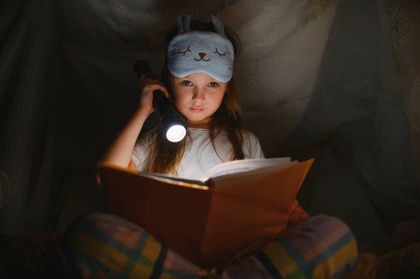 happy child girl laughing and reading book in dark in a tent at home - Фото, изображение