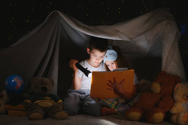 Two children with flashlight read a book under a blanket as a tent - Фото, зображення