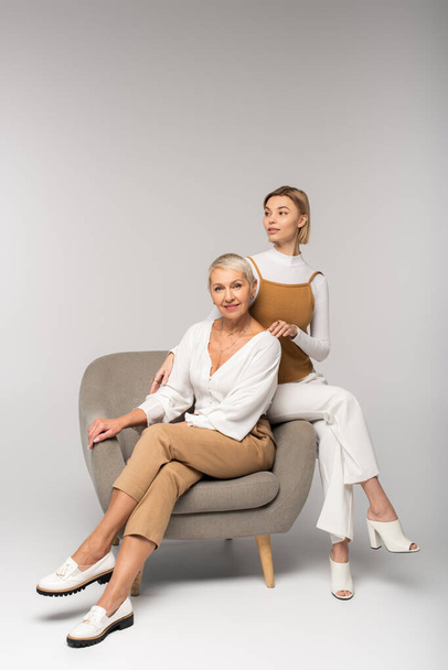 full length of young woman sitting near smiling middle aged mother in armchair on grey  - 写真・画像
