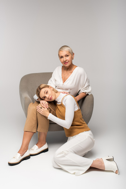 full length of young woman leaning on knees of mother sitting in armchair on grey  - Foto, Imagem