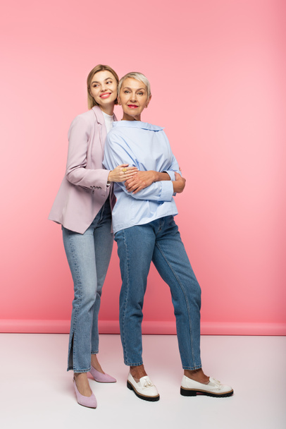 full length of happy young woman hugging mature mother in jeans and blouse while standing on pink - Fotografie, Obrázek
