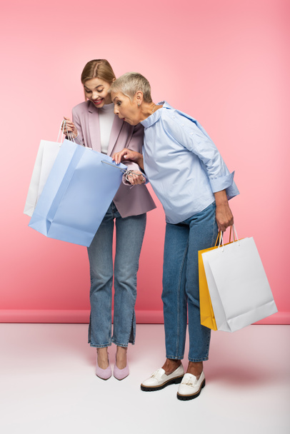 volledige lengte van vrolijke vrouw en verrast volwassen moeder in jeans op zoek naar shopping tassen op roze - Foto, afbeelding