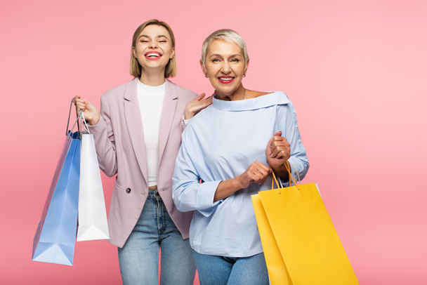 positive young woman and mature mother holding shopping bags isolated on pink  - Фото, зображення
