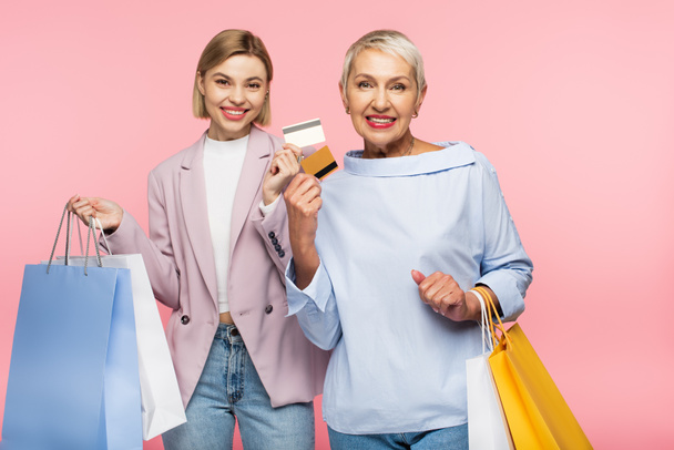 cheerful young woman and pleased mature mother holding shopping bags and credit cards isolated on pink  - Foto, Imagen