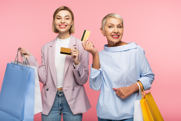 feliz joven y sonriente madre madura sosteniendo bolsas de compras y tarjetas de crédito aisladas en rosa  - Foto, Imagen
