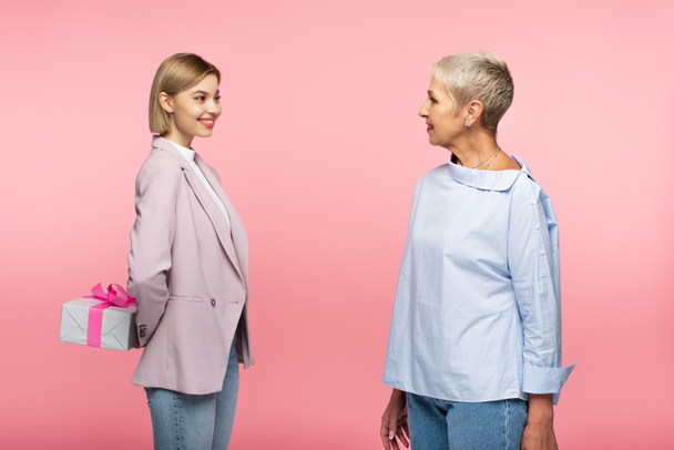 cheerful young woman holding present behind back near mature mother isolated on pink - Foto, afbeelding