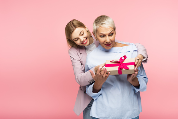 cheerful young daughter presenting gift to happy mother isolated on pink  - Photo, Image
