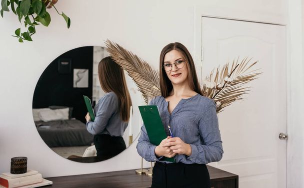 Porträt einer brünetten Frau mit Brille im Business-Stil, die mit Papieren im Raum steht und in die Kamera blickt - Foto, Bild