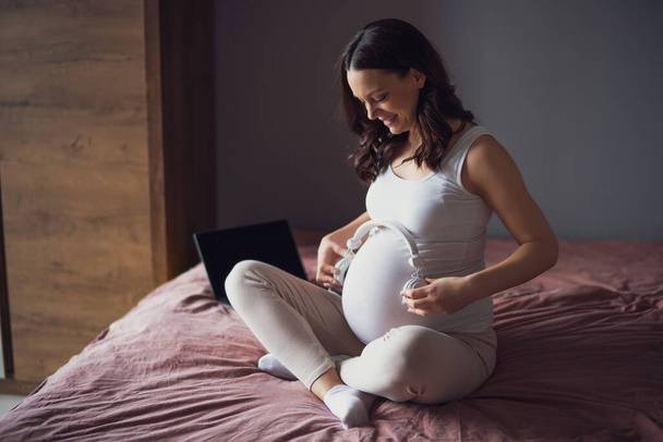 Feliz mujer embarazada relajándose en casa. Ella está sentada en la cama en el dormitorio y tocando música a su bebé en el estómago. - Foto, imagen