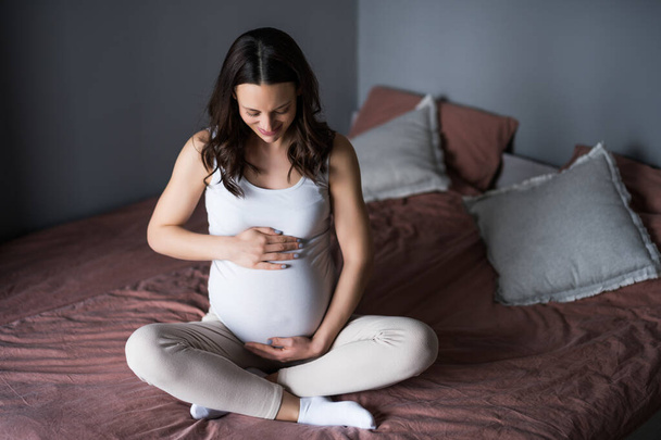 Happy pregnant woman relaxing at home. She is sitting on bed in bedroom. - Фото, зображення