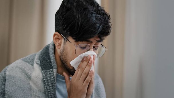 Close-up sad exhausted arabic bearded man suffering from seasonal allergy dust virus respiratory disease runny nose guy in glasses wrapped in blanket sitting at home blowing nose snot in paper napkin - Photo, Image