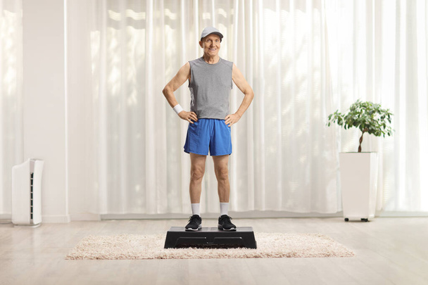 Full length portrait of an elderly man in sportswear standing on a stepper at home - Φωτογραφία, εικόνα