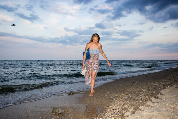 a young girl in the evening on the seashore in a sundress and with a hat, sunset, blue sky, clouds, calm, surf, sand, empty beach, wild - Foto, Imagen