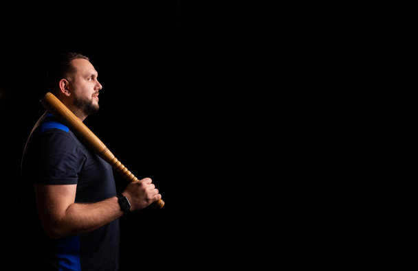 Young man with a baseball bat on a dark background. Bouncer, security guard, bodyguard concept. Place for text. A large man in a T-shirt stands sideways. - Foto, afbeelding
