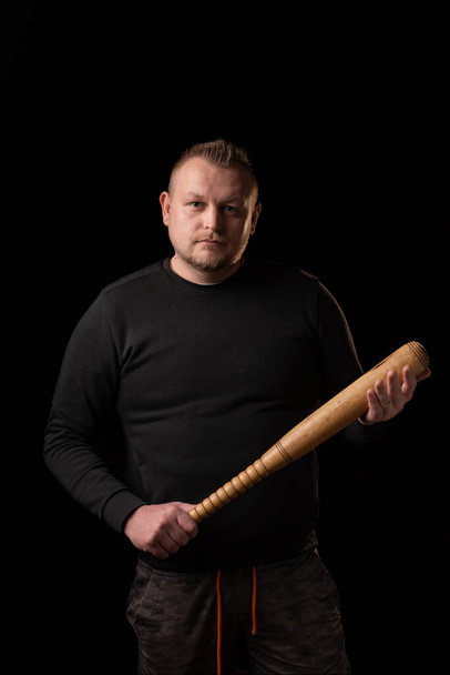 A young man is holding a wooden bat. Bouncer, security guard, bodyguard concept. A large man in a T-shirt looks at the camera. - Фото, изображение