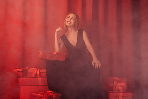The woman is sitting on a set of gifts. Middle-aged woman in a black dress. Red background, smoke. Happy face. Giving, receiving gifts concept. - 写真・画像