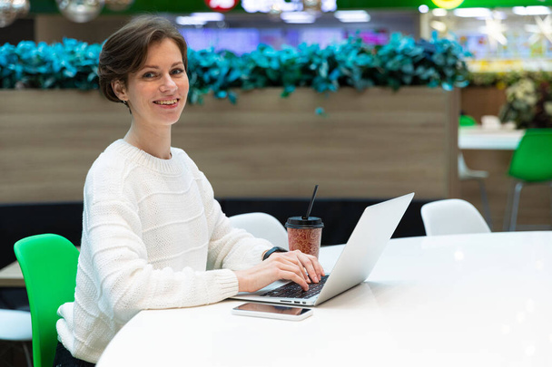 Photo of a young happy positive smiling woman who sits at the food court of a shopping center at a table and works at a computer laptop. Freelance concept - Foto, Imagen