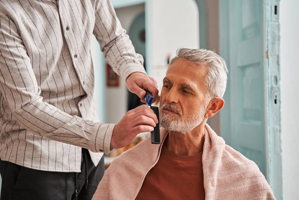 Grey haired man enjoying while his son cutting his beard with comb and trimer - Foto, Bild