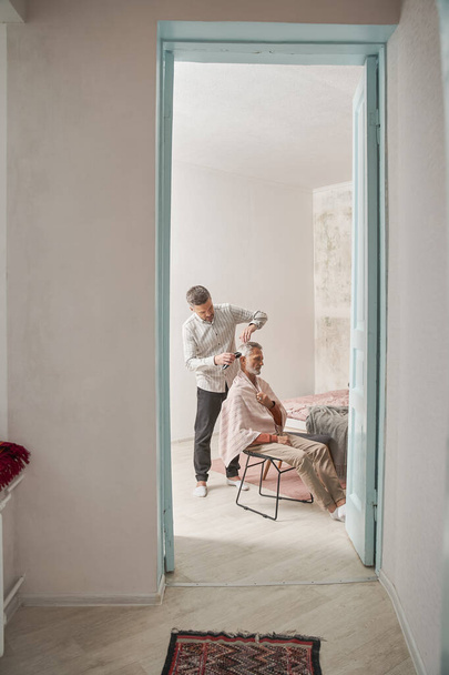 Hombre adulto cortando a su padre jubilado con trimmer mientras pasa tiempo en casa - Foto, imagen