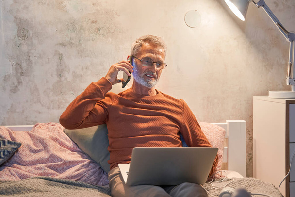 Smiling senior male in domestic clothes holding laptop and making phone call with smartphone - Foto, immagini