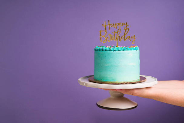 紫色の背景のコピースペースの前に金の幸せな誕生日サインを持つケーキを保持する女性の手 - 写真・画像