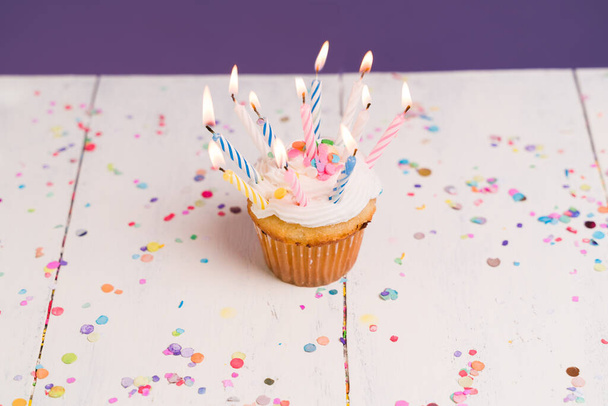 Geburtstagswunsch. Hoher Winkel eines leckeren Cupcake mit brennenden Geburtstagskerzen auf einem weißen Holztisch mit Konfetti - Foto, Bild