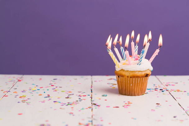 Many lit candles on a small delicious cupcake on a wooden table with colorful confetti in front of a purple background - Foto, afbeelding