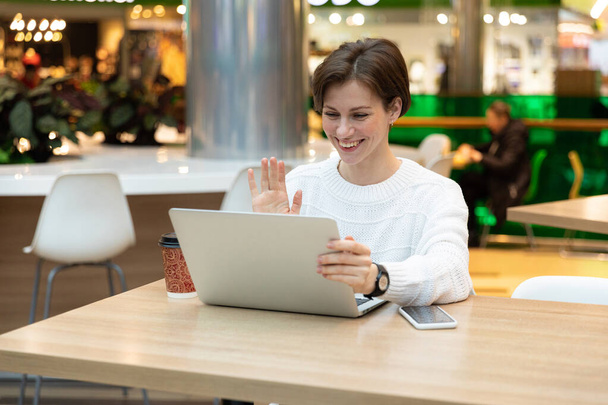 Junge positiv lächelnde schöne brünette Frau in weißem Sweatshirt sitzt in einem Einkaufszentrum an einem Tisch und arbeitet an einem Computer-Laptop. Freiberufler und Geschäftskonzept - Foto, Bild