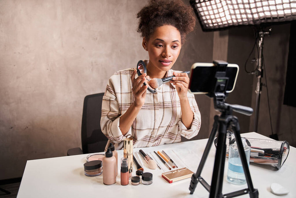 Multiracial woman picks up blush on the brush while broadcasting make up tutorial - Photo, image