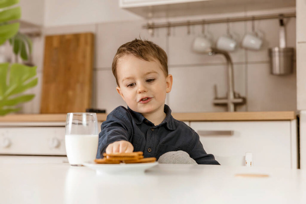 Little boy sitting in the kitchen and drinking milk. Fresh milk in glass, dairy healthy drink. Healthcare, source of calcium, lactose. Cozy and modern interior. Preschool child with casual clothing. - Фото, изображение