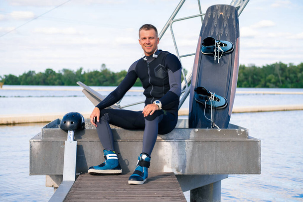 Wakeboarder confiant et souriant se reposant après l'entraînement, assis sur la jetée avec wakeboard - Photo, image