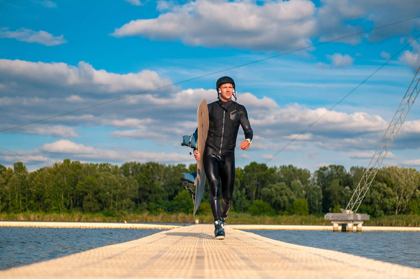 Hombre confiado en traje de neopreno con wakeboard caminando en puente flotante después de entrenar - Foto, Imagen