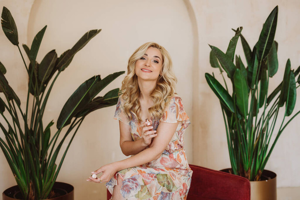 Sincere portrait of a beautiful blonde woman in a summer dress with a floral print on the background of large indoor plants in golden flowerpots. - Photo, Image