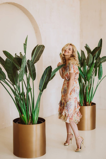 Piękna blondynka w letniej sukience z kwiatowym nadrukiem na tle dużych roślin Strelitzia w złotych doniczkach. - Zdjęcie, obraz