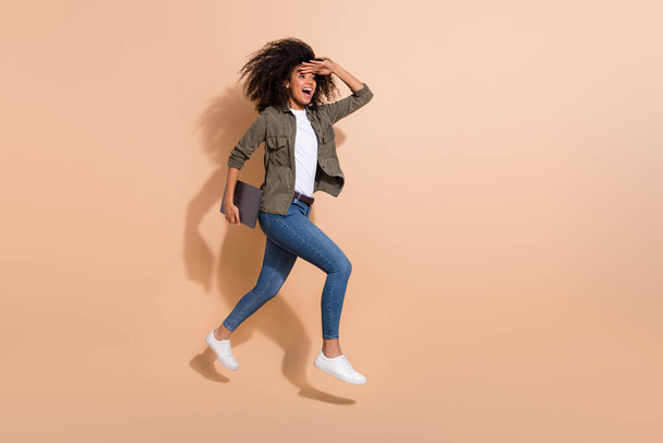 Full length body size view of pretty cheerful girl jumping carrying laptop finding way isolated over beige pastel color background - Foto, afbeelding