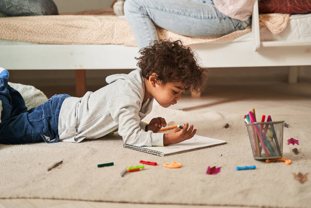 Little multiracial boy laying at the floor and drawing something with pencils at the paper - Photo, Image