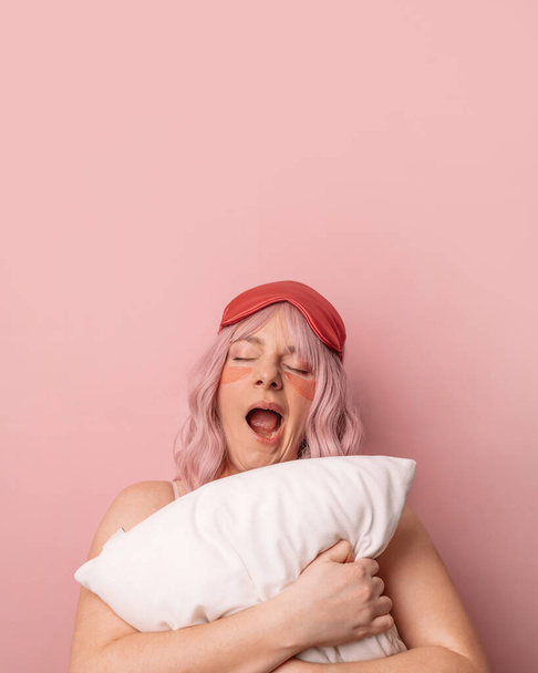 Красивая молодая довольно милая женщина носить маску сна держа мягкую подушку, зевает, как хочет спать, грустно просыпаться рано утром изолированы на розовом фоне. - Фото, изображение