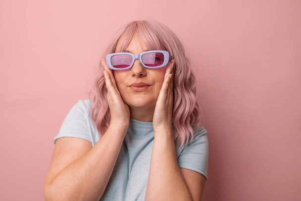 Feliz joven mujer caucásica en brillantes y elegantes gafas de sol de color magenta posando mirando a la cámara aislada sobre fondo rosa. - Foto, imagen