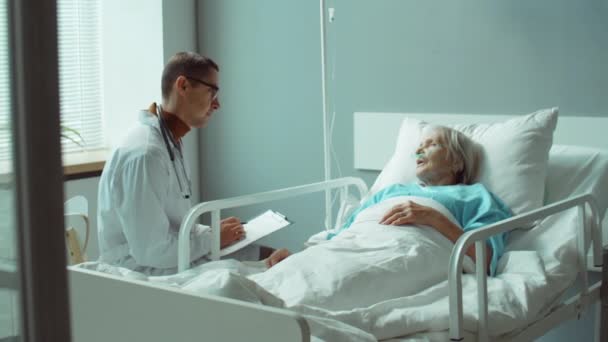 Επαγγελματίας άνδρας γιατρός με εργαστηριακή ποδιά κρατώντας πρόχειρο και έχοντας συζήτηση με την άρρωστη ηλικιωμένη γυναίκα, όπως αυτή βρίσκεται στο κρεβάτι στο θάλαμο του νοσοκομείου - Πλάνα, βίντεο