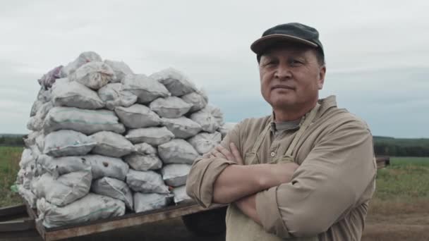 Horizontal médio retrato filmado de confiante maduro asiático agricultor de pé com braços cruzados contra sacos de batatas - Filmagem, Vídeo