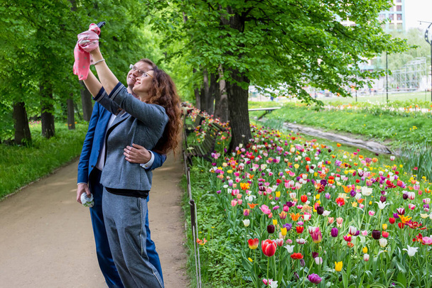 Moscú, Rusia, 13 de mayo de 2019: lindo joven y mujer con gafas que se toman fotos con un teléfono inteligente al aire libre en un día soleado de verano en el fondo de los tulipanes - Foto, Imagen