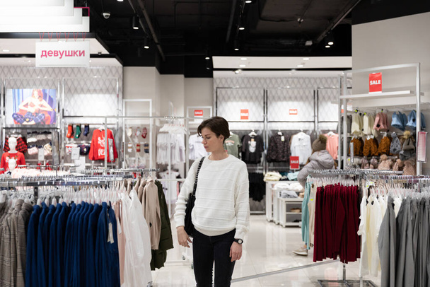 Een jonge mooie en aantrekkelijke brunette met kort haar in een casual en stijlvolle witte trui kiest modieuze kleding in een winkel in een winkelcentrum. Winkel interieur - Foto, afbeelding