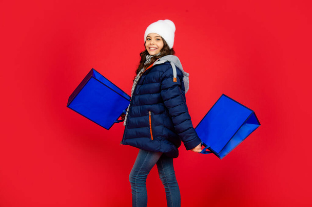 cheerful kid in puffer jacket and hat. teen girl after shopping on red background. - Foto, afbeelding