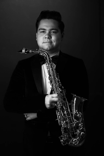 Black and White. Young Saxophonist in a Black Jacket, Hat and Sunglasses Holds a Baritone Saxophone. Dark Background. Neon Light. Close-up Portrait - Zdjęcie, obraz