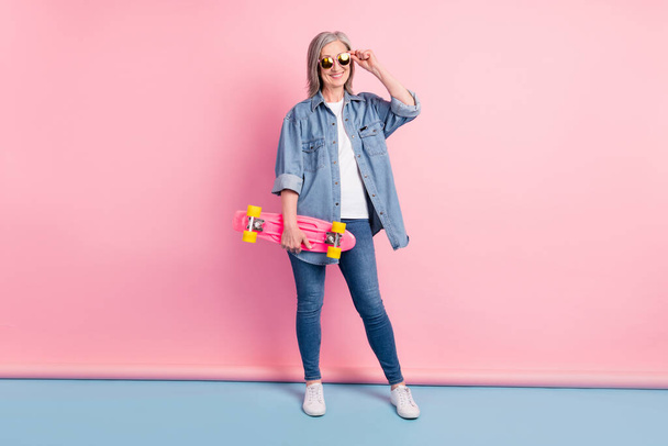 Full size photo of cool aged lady touch eyewear hold skate wear jeans shirt isolated on pink background - Foto, imagen