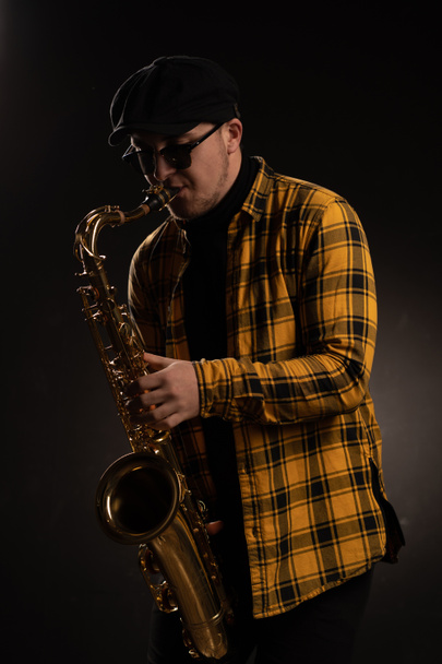 Man Saxophonist in Yellow Checkered Shirt, Cap and Stylish Sunglasses Holding an Alto Saxophone. Black Background. Close-up Portrait - 写真・画像